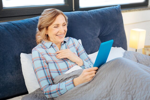 Regarder à l'écran. Dame adulte paisible se reposant dans son lit pendant la routine du matin et portant une tablette bleue dans les deux mains