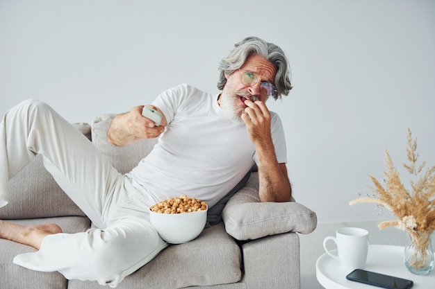 Regarder une émission de télévision et manger du pop-corn Senior homme moderne et élégant aux cheveux gris et à la barbe à l'intérieur