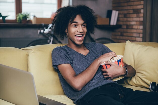 Regarder un film drôle. Gai jeune homme africain regardant la télévision et tenant un seau de pop-corn assis sur le canapé à la maison