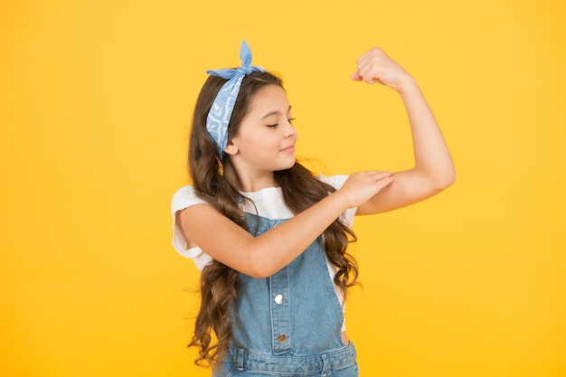 Regarder mes biceps confiants en puissance heureux après coiffeur petite beauté avec mouchoir cheveux bouclés enfant rétro fond jaune petite fille look vintage été enfant mode beauté et mode