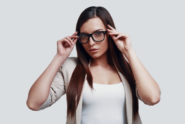 Regarder de près. Jolie jeune femme regardant la caméra et ajustant les lunettes en se tenant debout sur fond gris