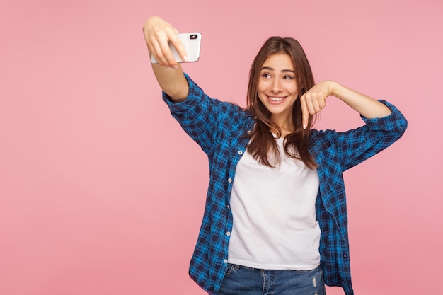 Regardez ci-dessous, abonnez-vous à la chaîne. Portrait d'une blogueuse heureuse en chemise à carreaux souriante et pointant vers le bas, en streaming vers les sous-marins, ayant une conversation positive en ligne. tourné en studio isolé sur fond rose