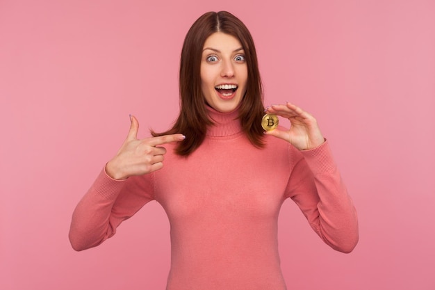 Regardez cette femme excitée surprise aux cheveux bruns en pull rose pointant du doigt le bitcoin en prêtant attention à la nouvelle crypto-monnaie numérique Prise de vue en studio intérieure isolée sur fond rose