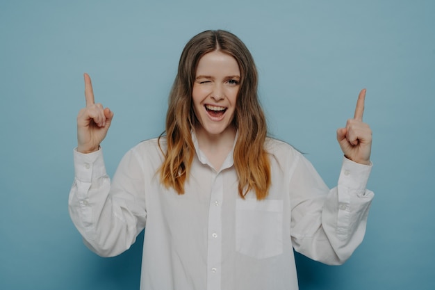 Regardez ici. Adolescente heureuse avec un large sourire en chemise blanche clignant de l'œil et pointant vers le haut avec ses doigts, debout isolée sur un mur bleu. Notion de publicité