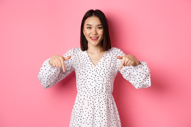 Regardez là. Jolie femme asiatique en robe, pointant les doigts vers le bas sur l'espace de copie, montrant une remise sur le produit et souriant, debout sur fond rose