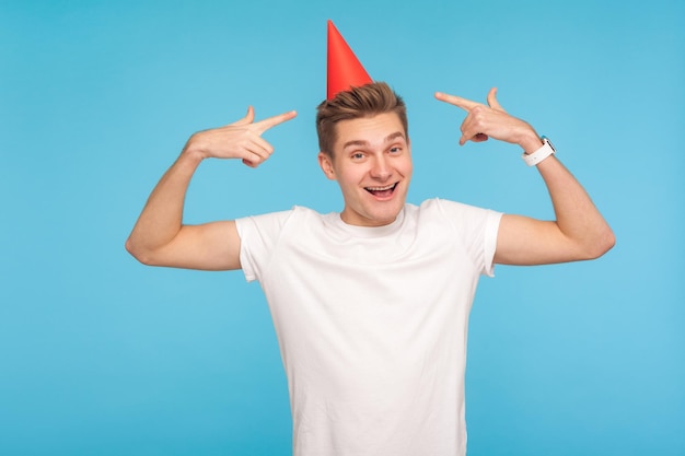 Regardez mon chapeau de fête. Portrait d'un homme joyeux en t-shirt pointant vers un drôle de cône sur sa tête et souriant à la caméra avec une expression insouciante enfantine. studio d'intérieur tourné isolé sur fond bleu