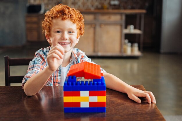 Regardez ce que j'ai construit. Adorable garçon aux cheveux bouclés assis à une table et s'excitant après avoir terminé son travail sur une maison en plastique colorée à la maison.