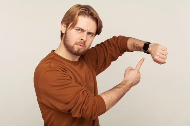 Regardez le temps. Portrait d'un homme impatient en colère avec une barbe en sweat-shirt pointant une montre-bracelet et ayant l'air ennuyé