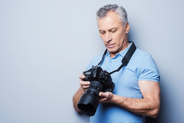 Réglage de la caméra. Portrait of senior man in T-shirt holding camera en se tenant debout sur fond gris