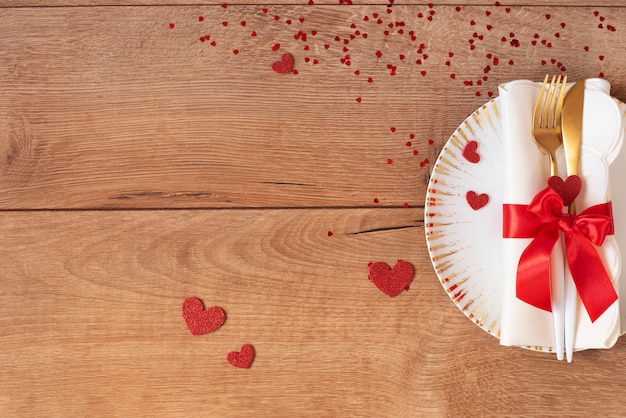 Réglage de la table de fête pour la Saint-Valentin avec fourchette, couteau, arc rouge et coeurs sur une table en bois. Espace pour le texte. Vue de dessus