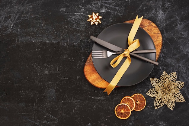 Réglage de la table noire avec une assiette sur une fourchette et un couteau de planche à découper avec des décorations de Noël à l'arc doré et des tranches d'orange Vue de dessus espace de copie à plat