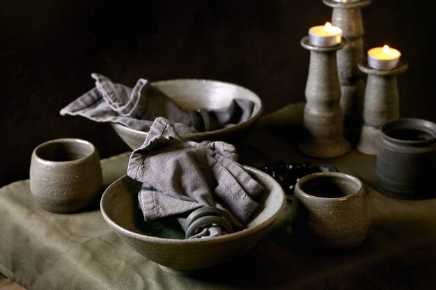 Réglage de la table rustique avec de la vaisselle en céramique artisanale vide, des bols et des tasses rugueux gris, des bougies allumées sur une nappe en lin. Fond sombre