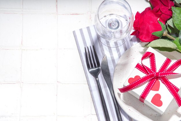 Réglage de la table de la saint-valentin avec boîte-cadeau assiette avec ruban rouge festif verre à vin fourchette et couteau roses rouges bouquet de fleurs table carrelée blanche flatlay copie espace