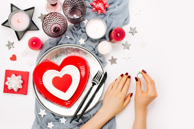 Réglage de la table de la Saint-Valentin. Les mains féminines tiennent la bague de mariage. Plats blancs et rouges en forme de coeur, verres à boisson, bougies et couverts sur fond blanc. Notion romantique