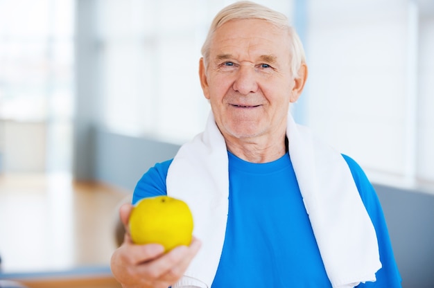 Rejoignez un mode de vie sain! Heureux homme senior avec une serviette sur les épaules qui s'étend de la pomme verte en se tenant debout dans un club de santé