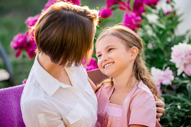 Des relations familiales heureuses. Maman et sa fille s'assoient sur un banc et discutent dans le jardin parmi les fleurs par une soirée d'été ensoleillée