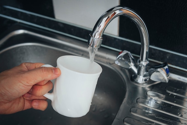 Remplissage du verre blanc avec de l'eau du robinet. Robinet et évier modernes dans la cuisine à domicile. Homme versant une boisson fraîche dans une tasse.