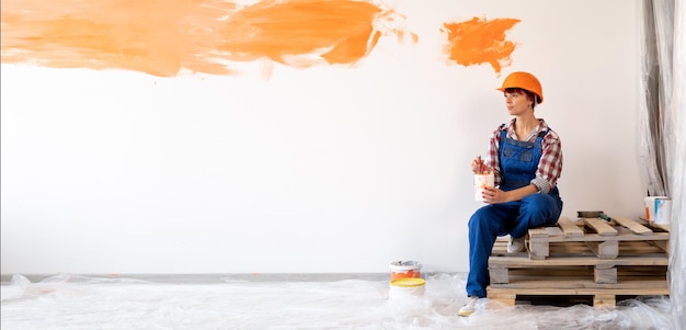 Photo rénovation de la maison et peinture murale une femme caucasienne avec des pinceaux à dessin dans un casque de protection et une combinaison est assise sur une palette en bois le concept d'appartements de réparation bannière