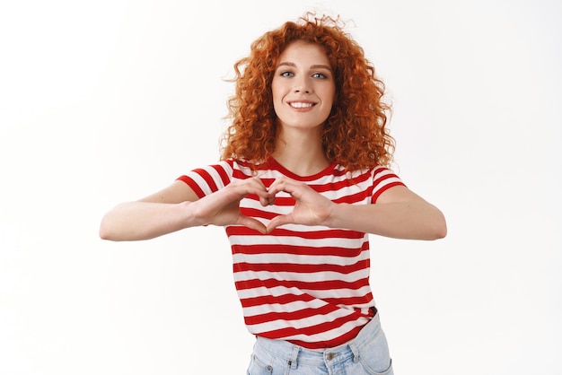 Répandre l'amour pas la guerre Attrayant positif heureux souriant européen rousse fille bouclée 25 ans portant un t-shirt rayé montrer signe de coeur geste souriant adorer exprimer affection sympathie fond blanc