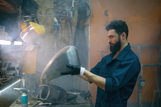 Réparateur concentré sérieux inspectant le nouveau réservoir d'essence soudé pour moto. Militaire travaillant dans un atelier enfumé le soir