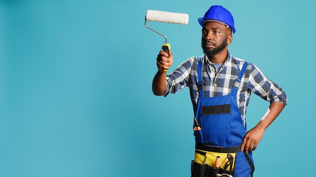 Réparateur en salopette peignant les murs avec une brosse à rouleaux, utilisant un équipement de rénovation devant la caméra. Charpentier confiant avec casque travaillant avec un pinceau roulant pour changer de couleur en studio.