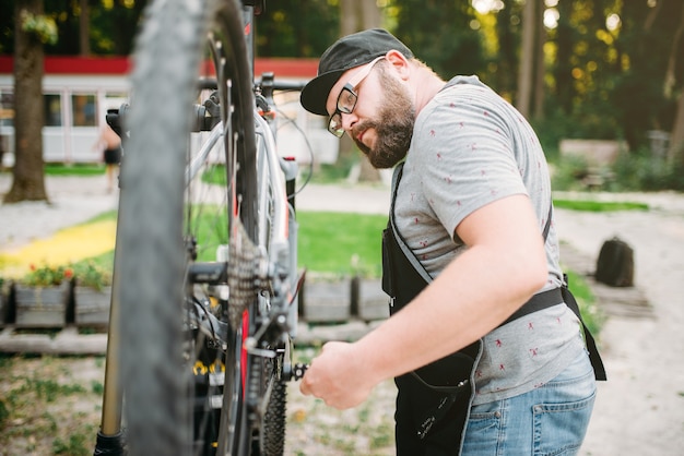 Réparateur travaille avec roue de vélo, atelier de vélo en plein air. Mécanicien vélo barbu en tablier