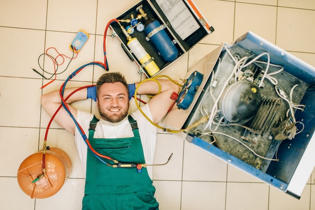 Réparateur en uniforme se trouve près du réfrigérateur à la maison, vue de dessus. Réparation d'occupation de frigo, service professionnel