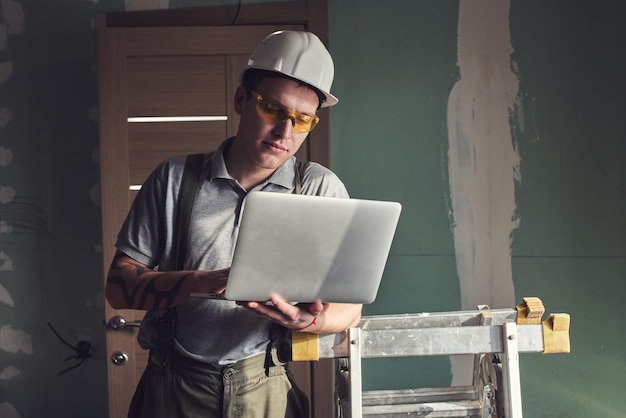 Réparation de chambre. Constructeur en casque et lunettes se tient avec un ordinateur portable dans ses mains sur le fond de la construction