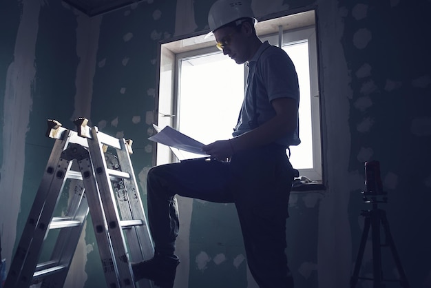 Réparation de chambre. Constructeur en casque et lunettes se tient avec un ordinateur portable et des dessins dans ses mains sur le fond de la construction