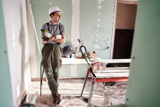 Réparation de chambre. Jeune constructeur dans un casque et une salopette debout avec les mains jointes sur le fond du chantier de construction