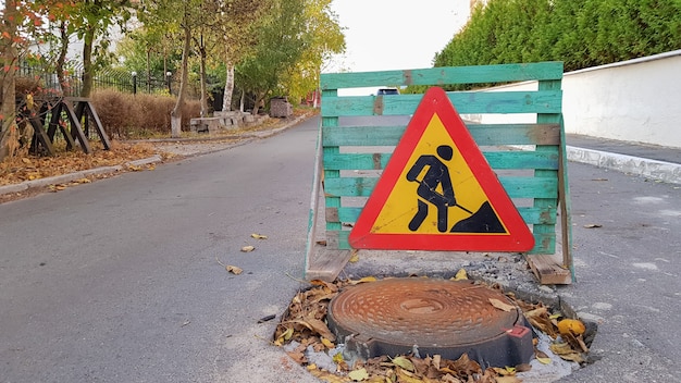 Réparation De Routes. Regard D'égout Sur La Chaussée. Les Conducteurs De Voitures D'avertissement Triangulaires Signent Avec Un Homme Avec Une Pelle Jaune Avec Du Rouge. Hommes Au Travail. Panneaux De Circulation, Restrictions De Voyage.