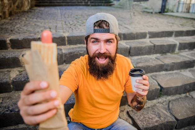 Repas qui a bon goût. Homme barbu mangeant des aliments malsains pendant le repos et la pause repas. Hipster mangeant un repas de hot-dog dans les escaliers en plein air. Un gars de race blanche aime manger des plats à emporter.