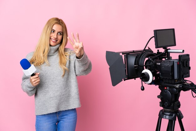 Reporter femme tenant un microphone et rapportant des nouvelles sur un mur rose isolé heureux et compter trois avec les doigts