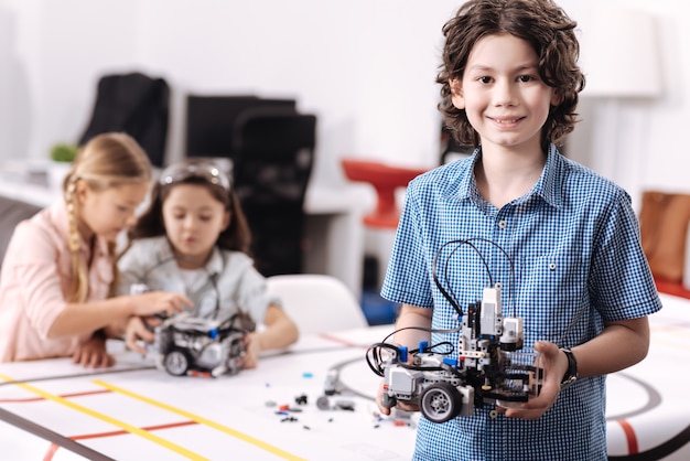 Représentant notre projet. Sourire heureux joyeux garçon debout à l'école et tenant un robot pendant que ses camarades de classe travaillent sur le projet