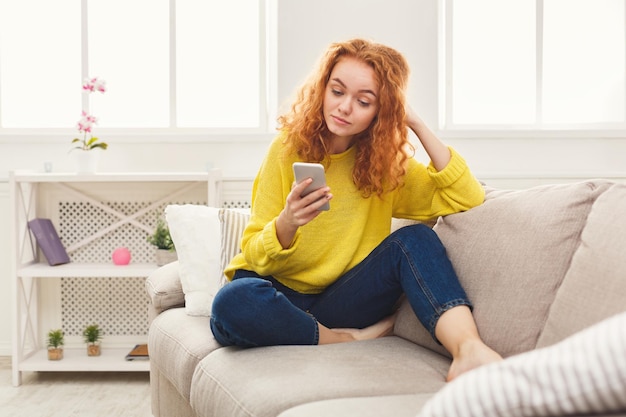 Réseaux sociaux. Jeune femme rousse envoyant des messages sur un smartphone à la maison, assise confortablement sur un canapé beige, espace pour copie