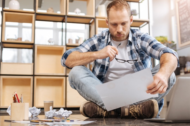 Respect de la date limite. Pensive fatigué homme travaillant dur assis les jambes croisées sur le sol et tenant la feuille de papier tout en travaillant sur le projet