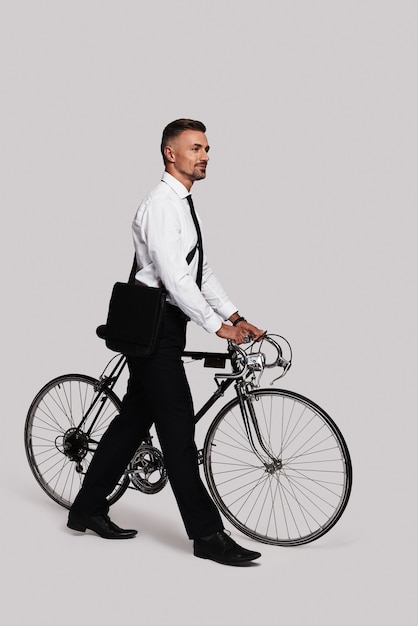 Respectueux de l'environnement. Toute la longueur du beau jeune homme en costume complet tirant son vélo et souriant en marchant sur fond gris