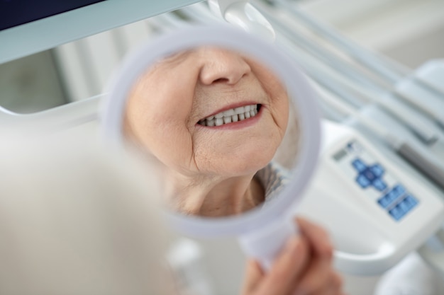 Restauration des dents. Femme aînée regardant dans le miroir après avoir visité le bureau de dentistes