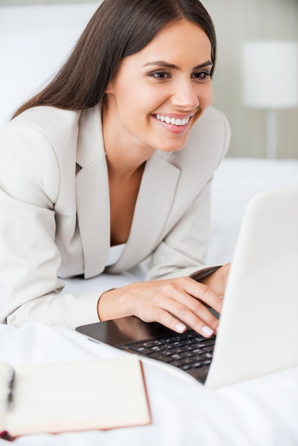 Rester en contact avec les collègues au bureau. Jeune femme d'affaires confiante en costume travaillant sur un ordinateur portable et souriante en position couchée dans son lit dans la chambre d'hôtel