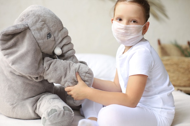 Restez à la maison en quarantaine prévention de la pandémie de coronavirus. Enfant triste en masque médical de protection et son éléphant tous deux en masque médical de protection assis sur le lit