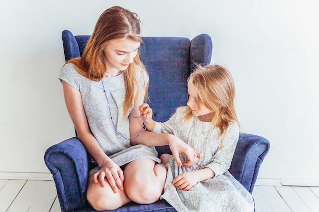 Restez à La Maison Restez En Sécurité. Deux Enfants Heureux Assis Sur Une Chaise Bleue Confortable Relaxant Jouant Dans Le Salon Blanc à L'intérieur. Petite Fille Jouant Avec Une Adolescente Montrant Ses Soins Amoureux. Sœurs S'amusant à La Maison.