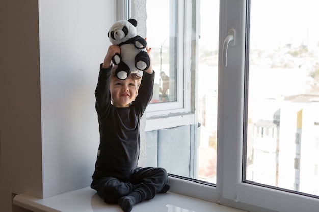 Restez positif à la maison. heureux et gai Un enfant est assis sur un rebord de fenêtre et joue avec un ours en peluche panda
