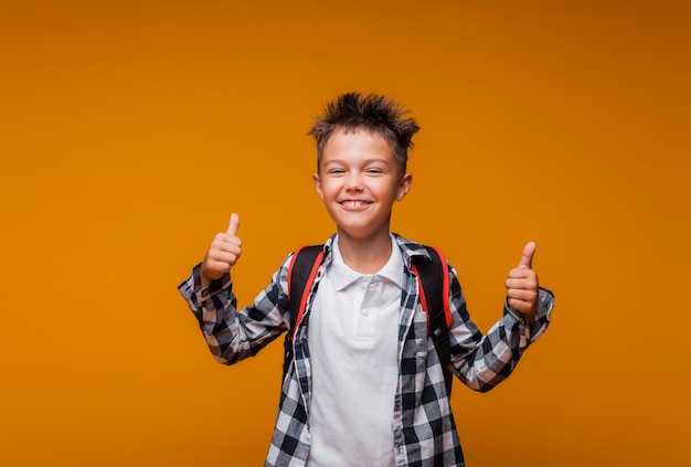 Photo de retour à l'école, un garçon drôle avec un sac à dos dans une chemise sur fond jaune montre une super classe et des sourires regarde l'espace de la caméra pour copier