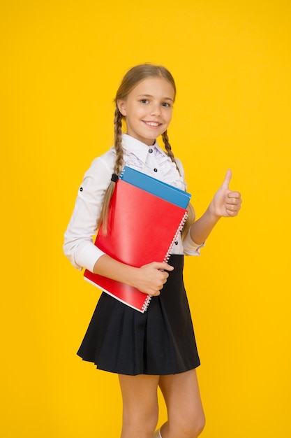 Retour à l'école. petite fille en uniforme montre le pouce vers le haut. enfant après les cours portent des documents. étudier à la maison. style de mode scolaire. fond jaune étudiant à la recherche intelligente. concept d'éducation moderne.