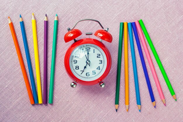 Retour à l'école. Réveil rouge et crayons de couleur sur fond violet, vue de dessus.