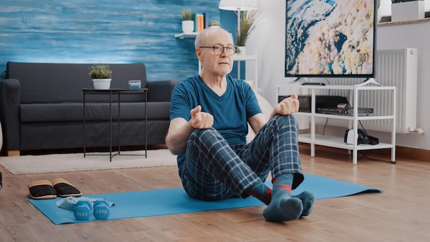 Retraité calme en position du lotus faisant de la méditation pour se détendre sur un tapis de yoga. Homme senior méditant pour pratiquer le bien-être et la relaxation. Personne âgée faisant des activités de Pilates pour l'harmonie.