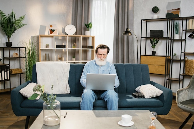 Retraité caucasien avec barbe grise en tenue décontractée assis sur un canapé confortable et travaillant sur un ordinateur portable