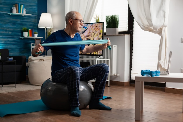 Retraité senior man sitting on swiss ball in living room faisant des exercices de remise en forme de soins de santé