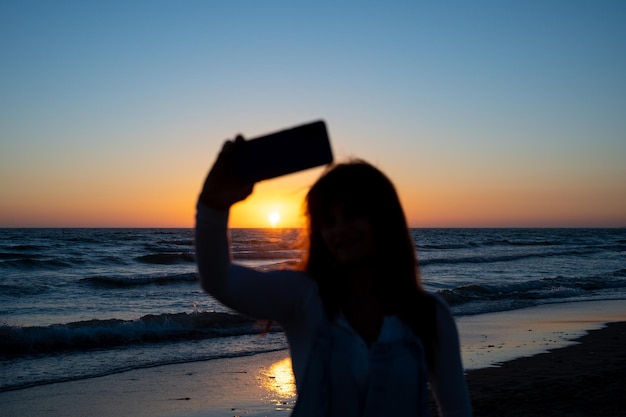 Rétro-éclairage d'une femme méconnaissable prenant un selfie d'un coucher de soleil en mer