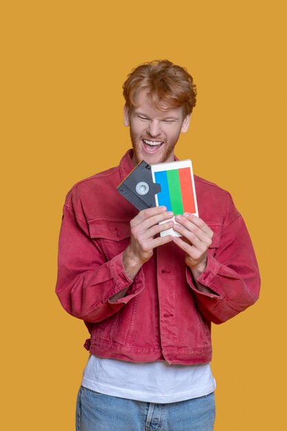 Rétro. Jeune homme barbu aux cheveux roux en veste rouge tenant une cassette vidéo